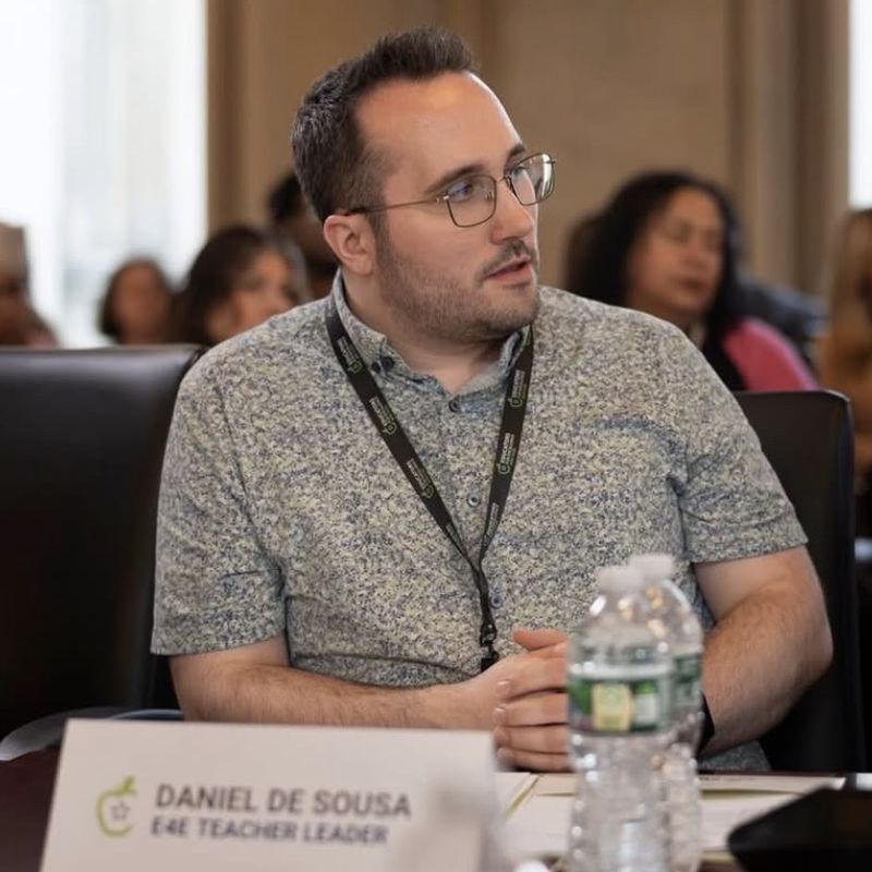 Dan speaking at a meeting table behind a name card that reads Daniel De Sousa, E4E Teacher Leader.